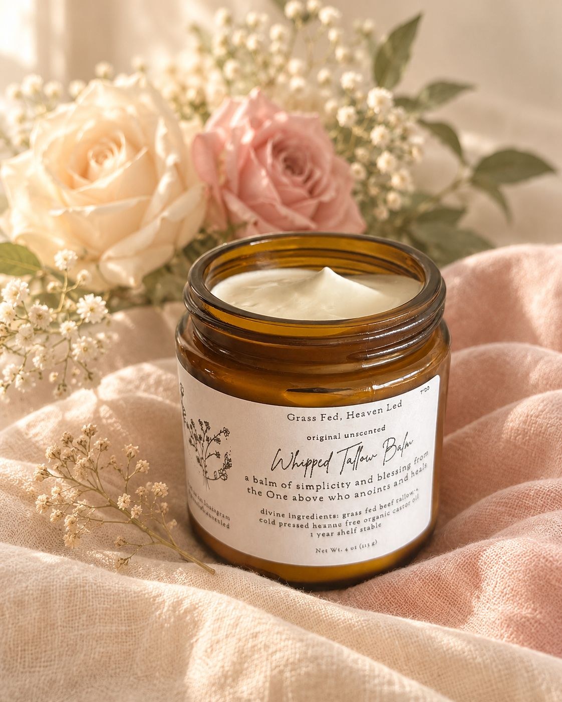 An open amber jar of Grass Fed, Heaven Led whipped tallow balm resting on soft blush linen beside cream and pink roses